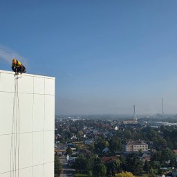 LT Labocatech Usługi wysokościowe, usługi alpinistyczne - Pracownik w kasku i uprzęży na krawędzi wysokiego budynku z białymi panelami, przygotowujący się do pracy na wysokości. W tle panorama miasta z mostem i kominem.