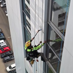 LT Labocatech Usługi wysokościowe, usługi alpinistyczne - Pracownik w uprzęży, z żółtym kaskiem, czyści okna na wysokościowcu za pomocą szczotki na teleskopowym kiju. Widok z góry na ulicę z zaparkowanymi autami.