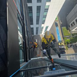 LT Labocatech Usługi wysokościowe, usługi alpinistyczne - Alpiniści przemysłowi w kaskach czyszczą szklaną fasadę biurowca za pomocą szczotek teleskopowych. Widok z góry na budynek i parking.