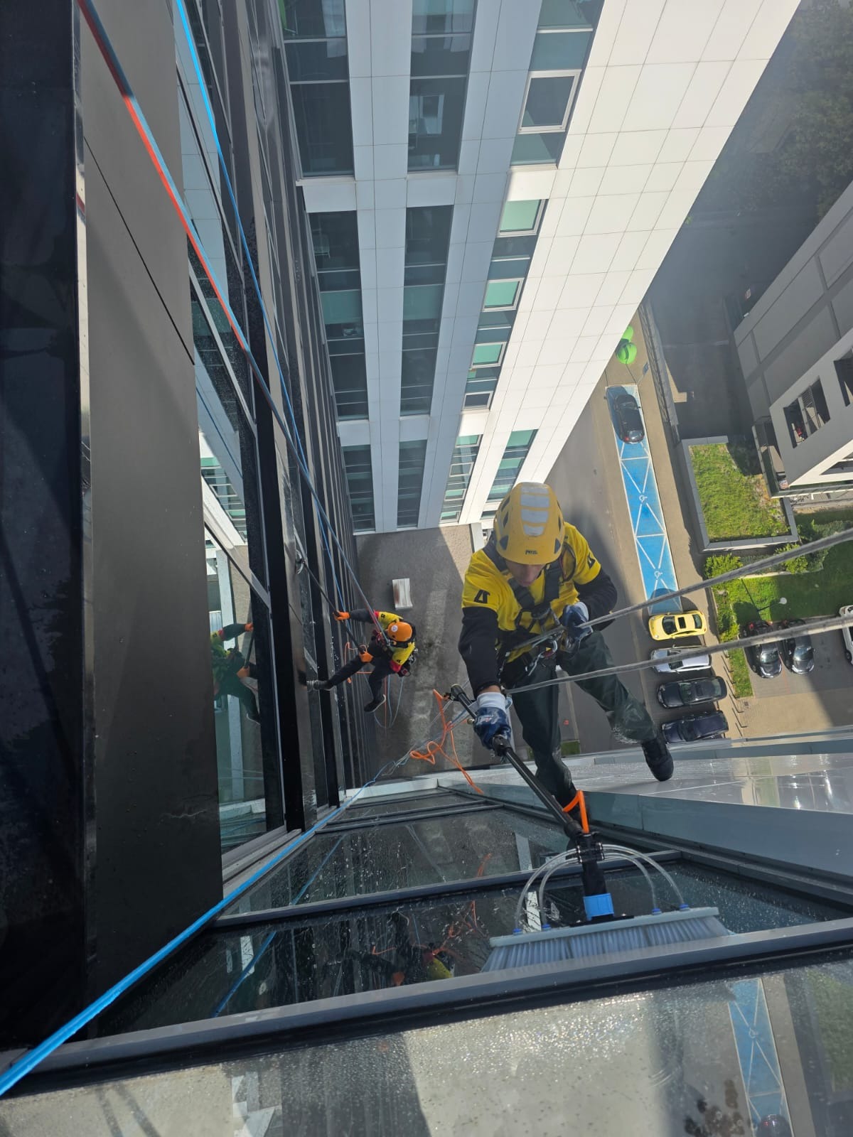 Dwóch alpinistów w kaskach i uprzężach czyści szklaną fasadę budynku za pomocą specjalistycznego sprzętu. Widok z góry, z perspektywy liny.