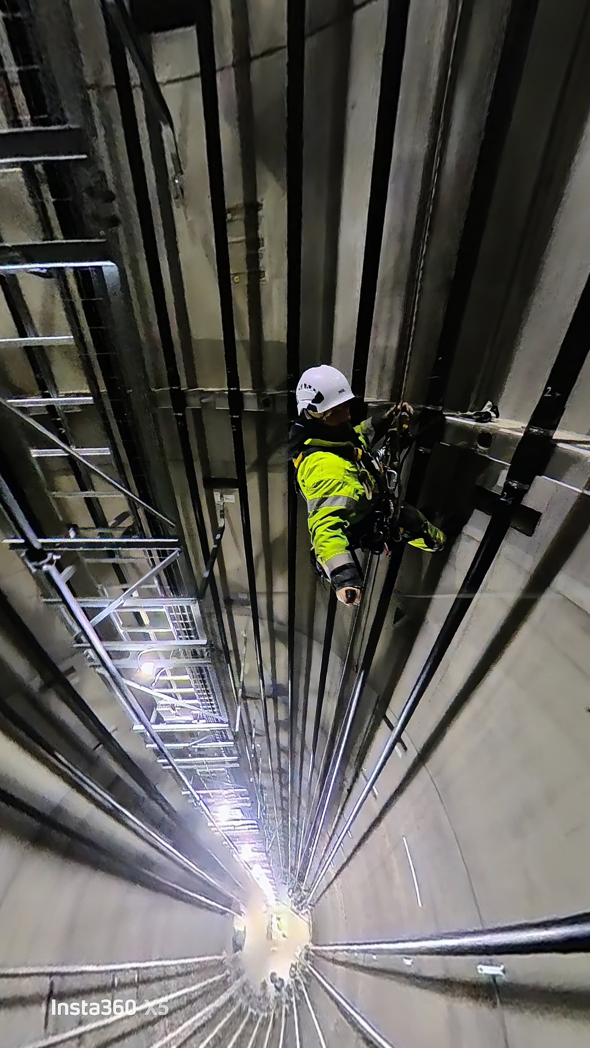Pracownik w kasku i jaskrawej kurtce, zabezpieczony linami, w szybie windy. Widok z dołu, perspektywa zniekształcona, oświetlenie surowe, metalowe elementy konstrukcyjne.