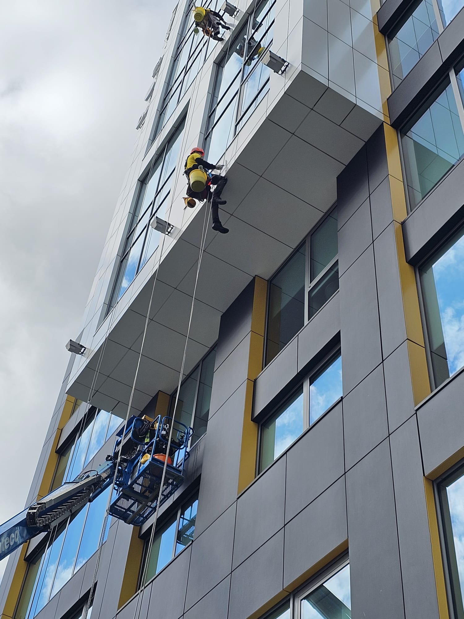 Pracownicy alpinistyczni podczas czyszczenia szklanej fasady biurowca z użyciem podnośnika koszowego i technik linowych. Widok z dołu na budynek.