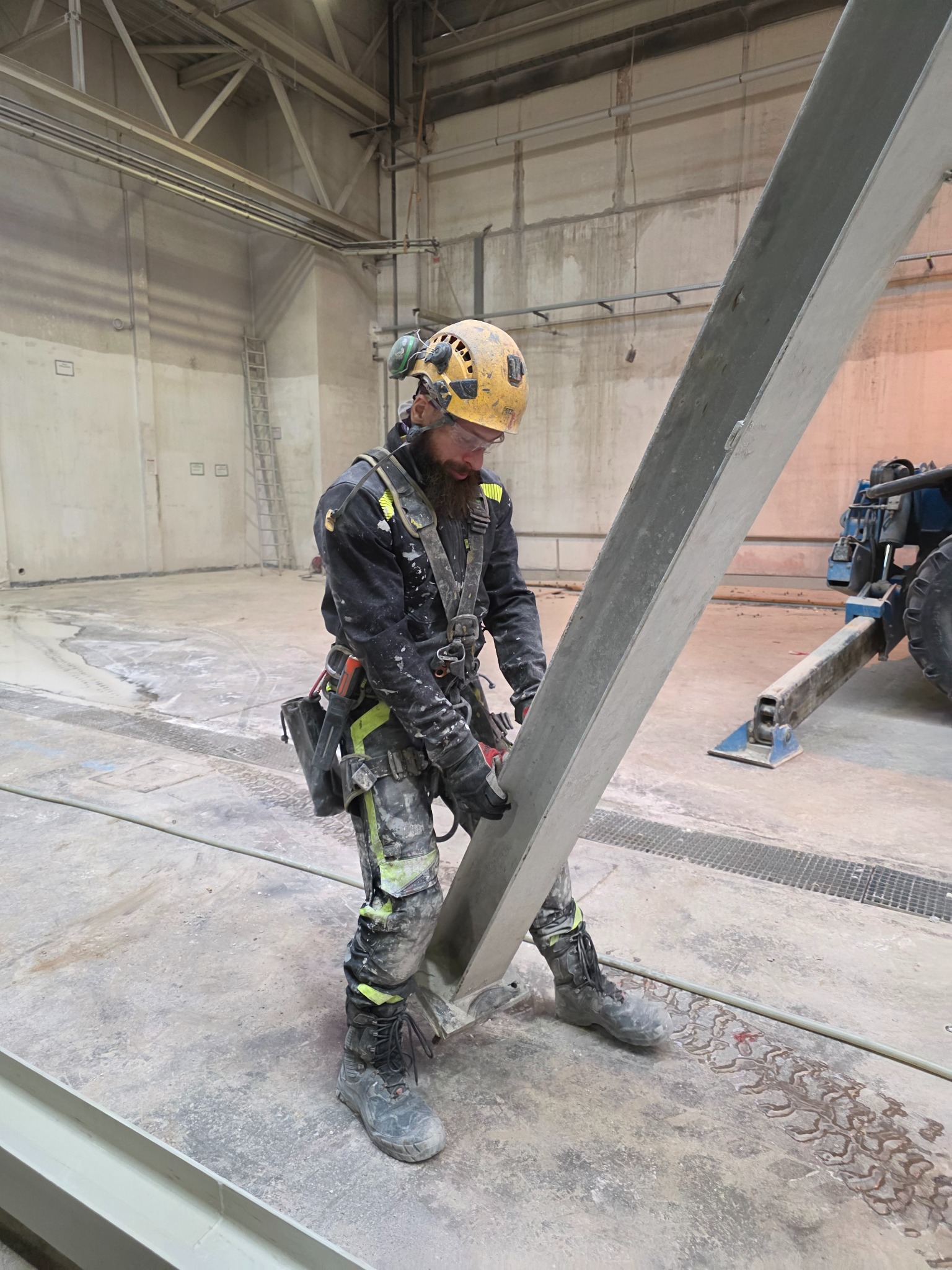 Pracownik w kasku i uprzęży, w trakcie prac wysokościowych wewnątrz hali, trzymający metalowy element konstrukcyjny. W tle widoczny podnośnik i drabina.