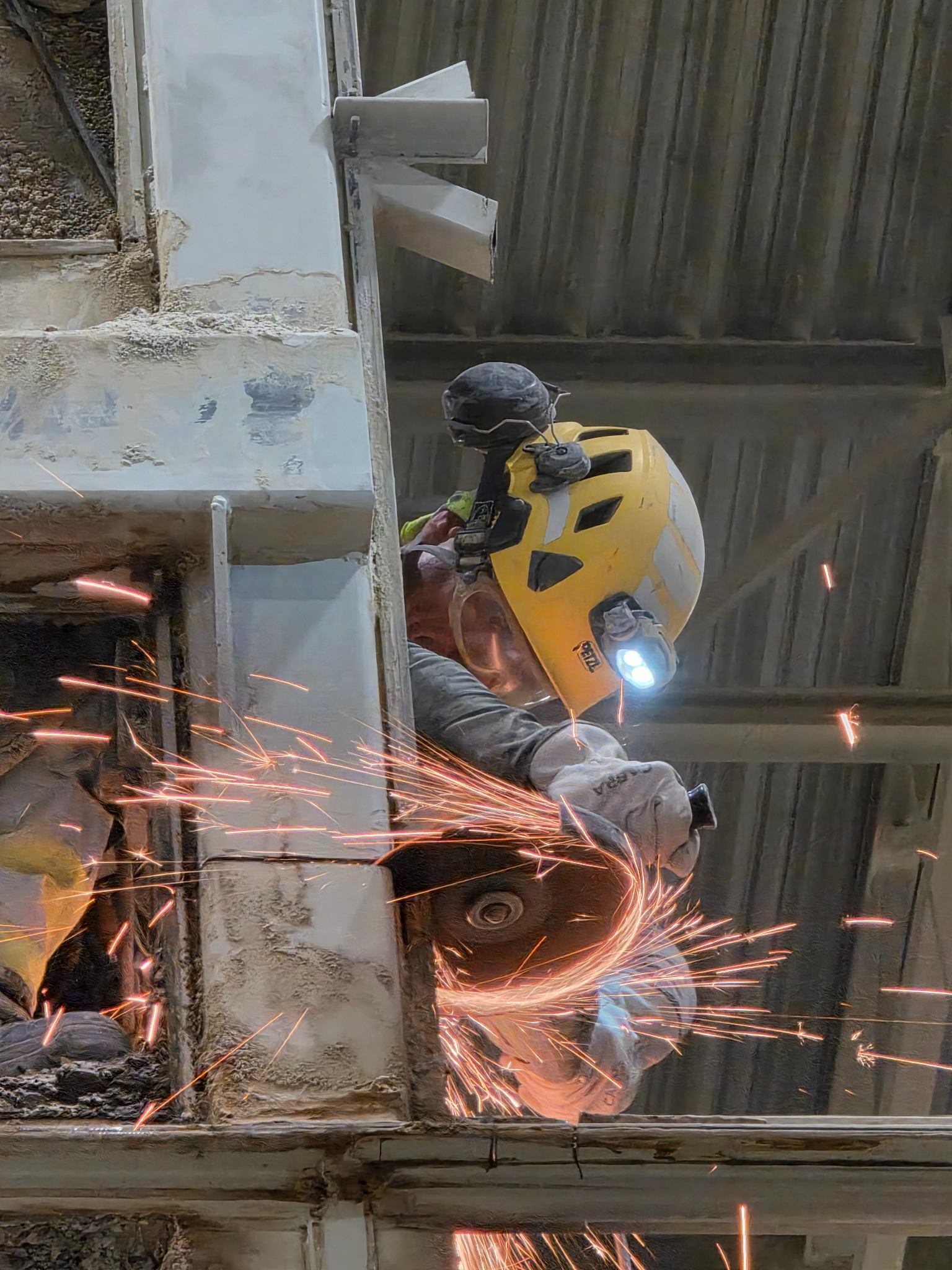 Pracownik w kasku tnie metalową konstrukcję szlifierką kątową, iskry rozświetlają otoczenie. Widoczne elementy konstrukcji i fragment dłoni w rękawicy.