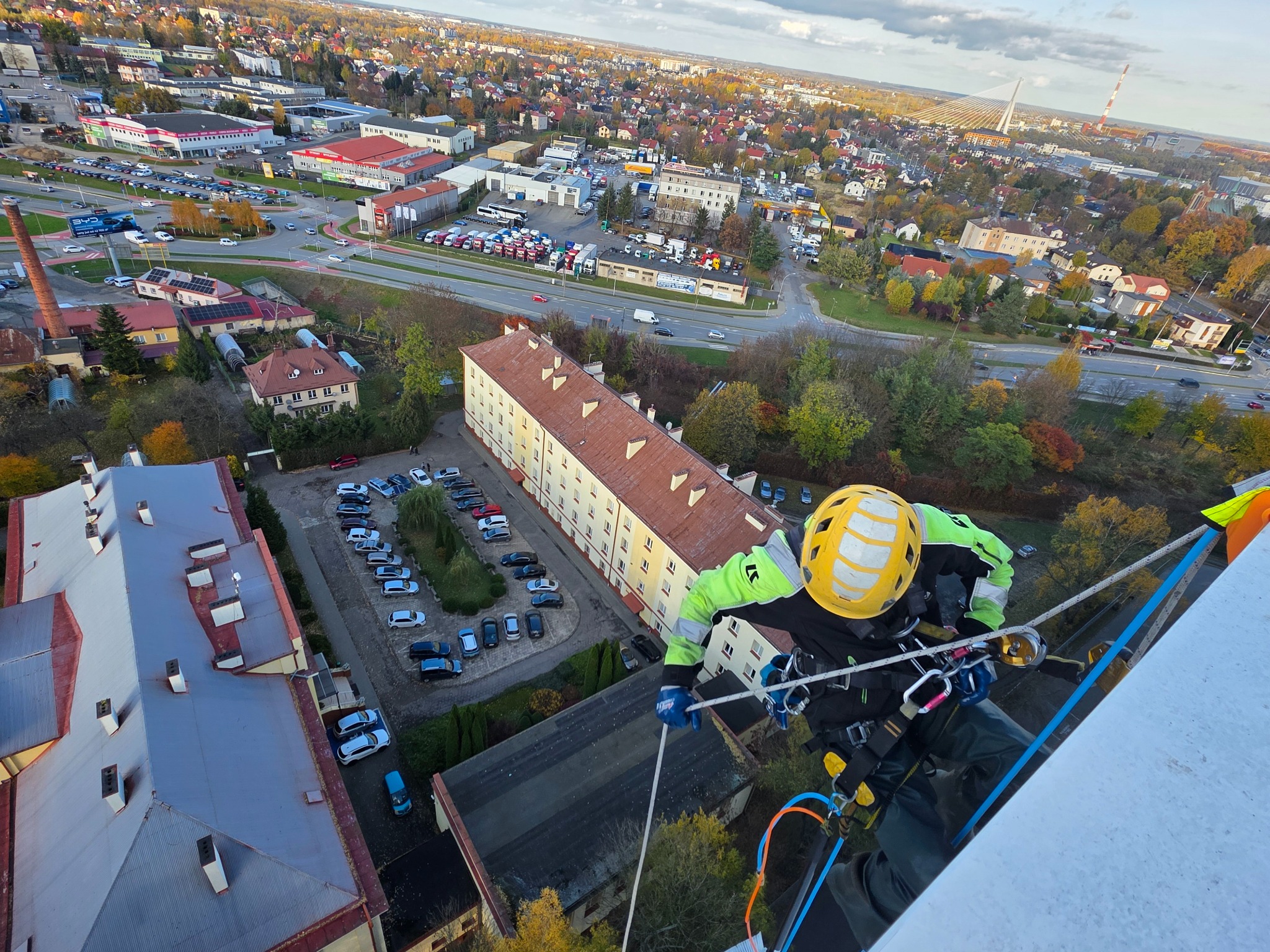 Pracownik w kasku i uprzęży na wysokości, z widokiem na miasto. Ujęcie z góry, z perspektywy osoby wykonującej prace wysokościowe na budynku.