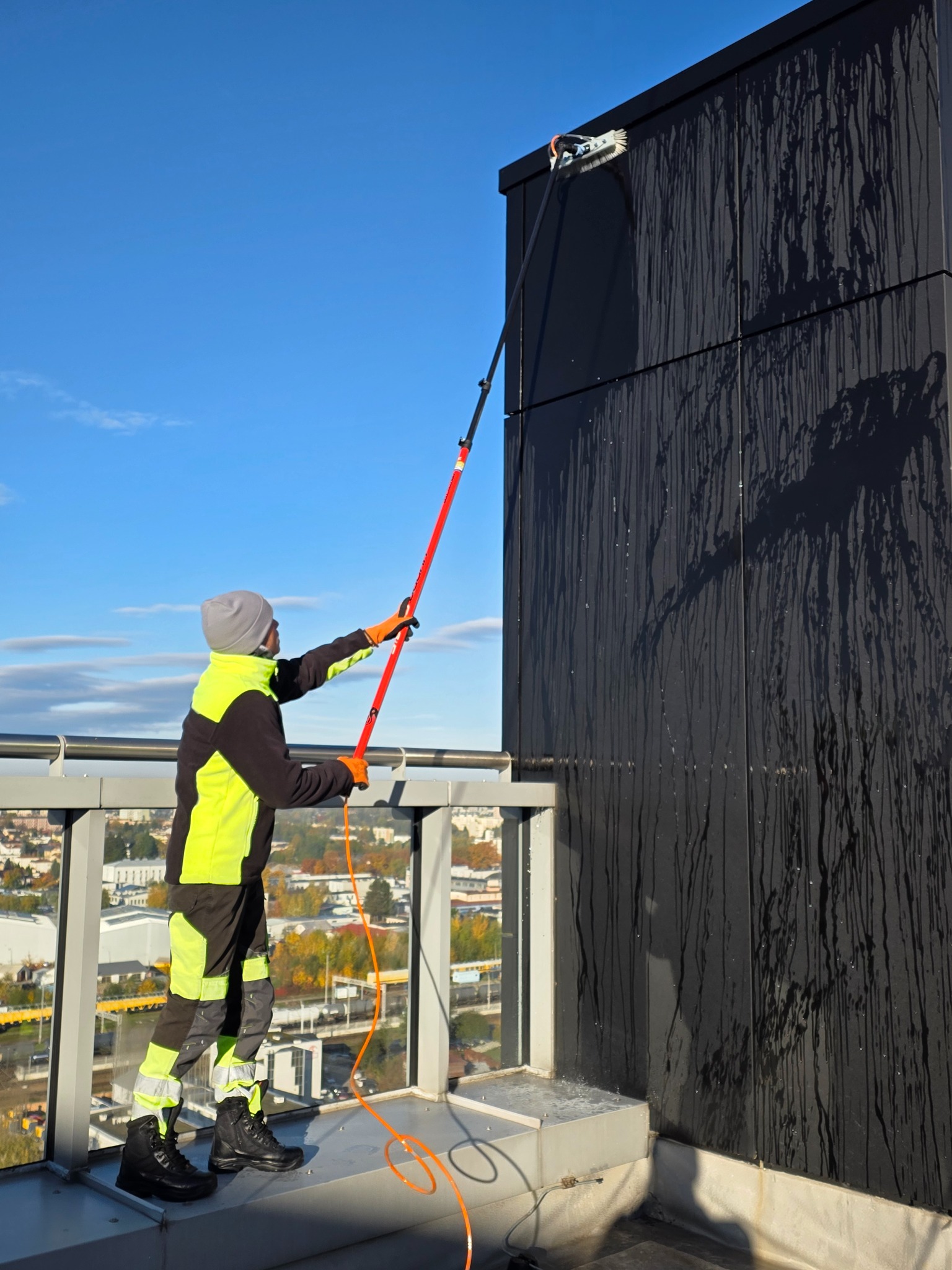 Pracownik w odzieży ochronnej myje ciemną elewację budynku za pomocą szczotki na długim wysięgniku. Widoczne zacieki i panorama miasta w tle.