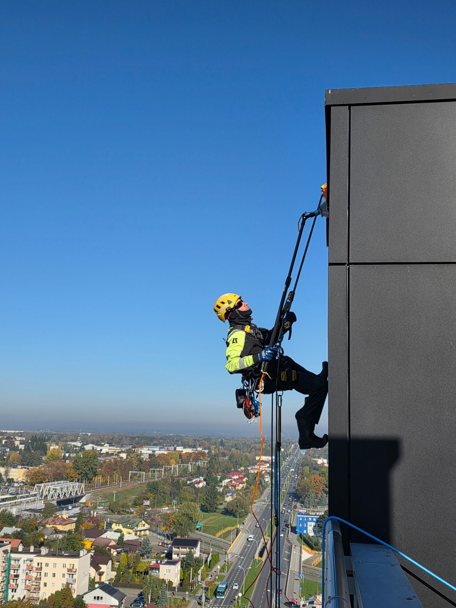 Pracownik w kasku, zabezpieczony linami, czyści elewację budynku z użyciem specjalistycznego sprzętu. Widok z góry na miasto w słoneczny dzień.