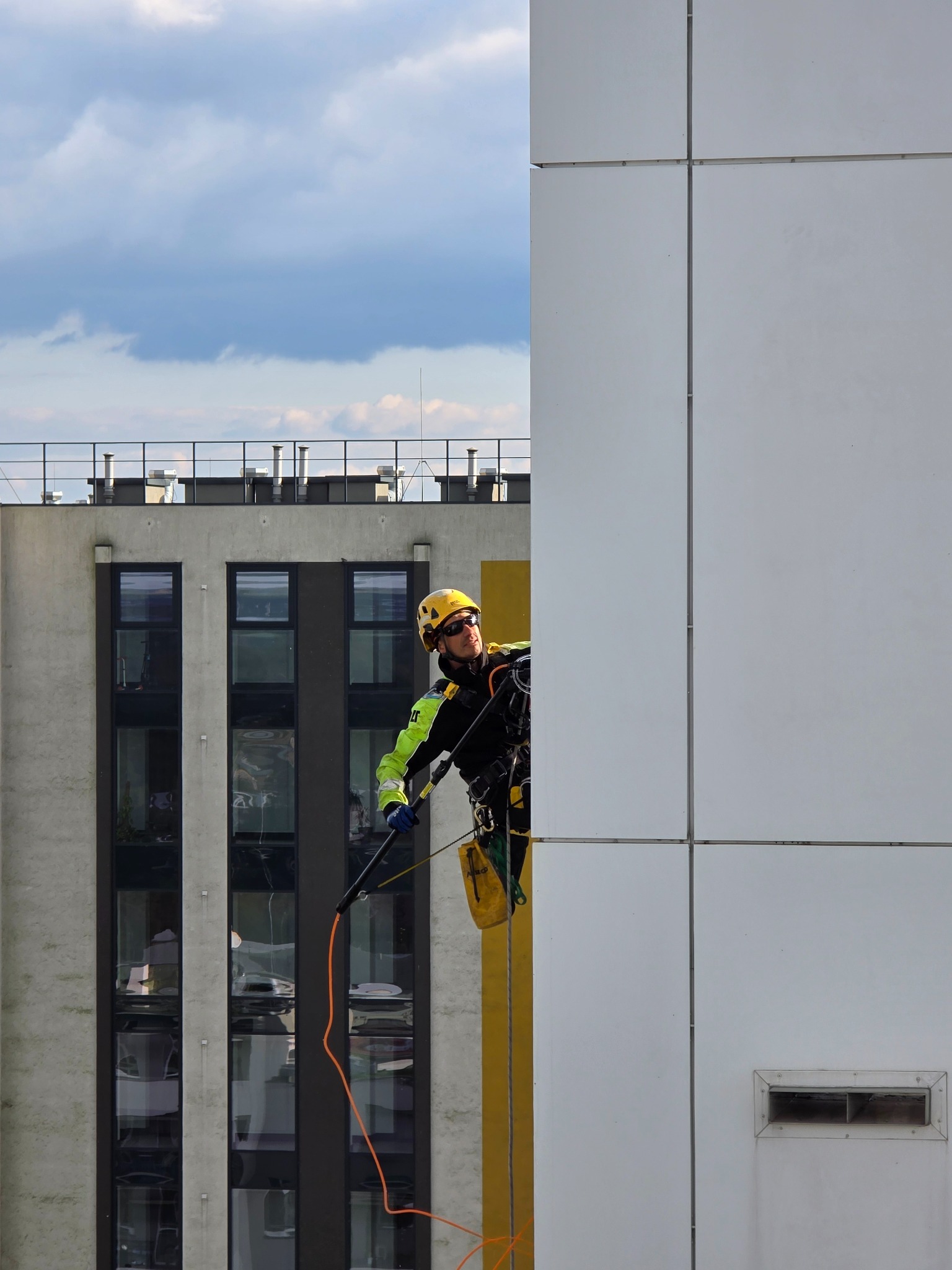 Pracownik w uprzęży, z lancą, czyści elewację budynku z białych paneli. Widoczne okna budynku w tle i fragment nieba.