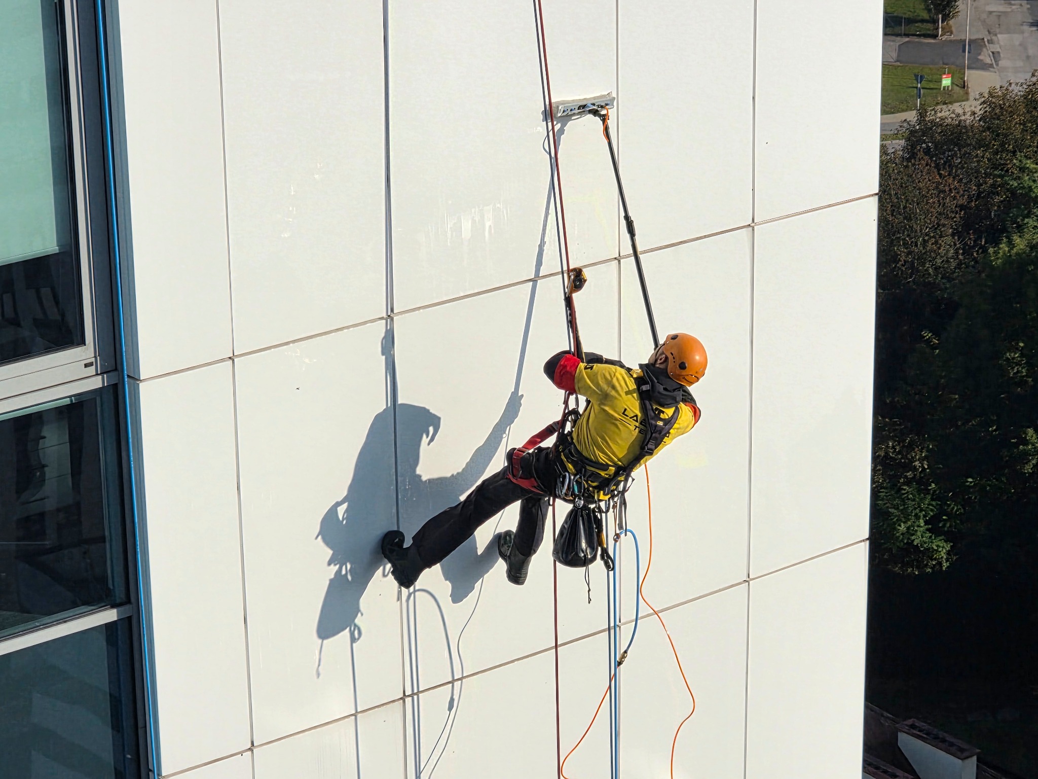 Pracownik w kasku, na linach, myje białą elewację budynku za pomocą specjalistycznego sprzętu. Widok z góry, słoneczny dzień.