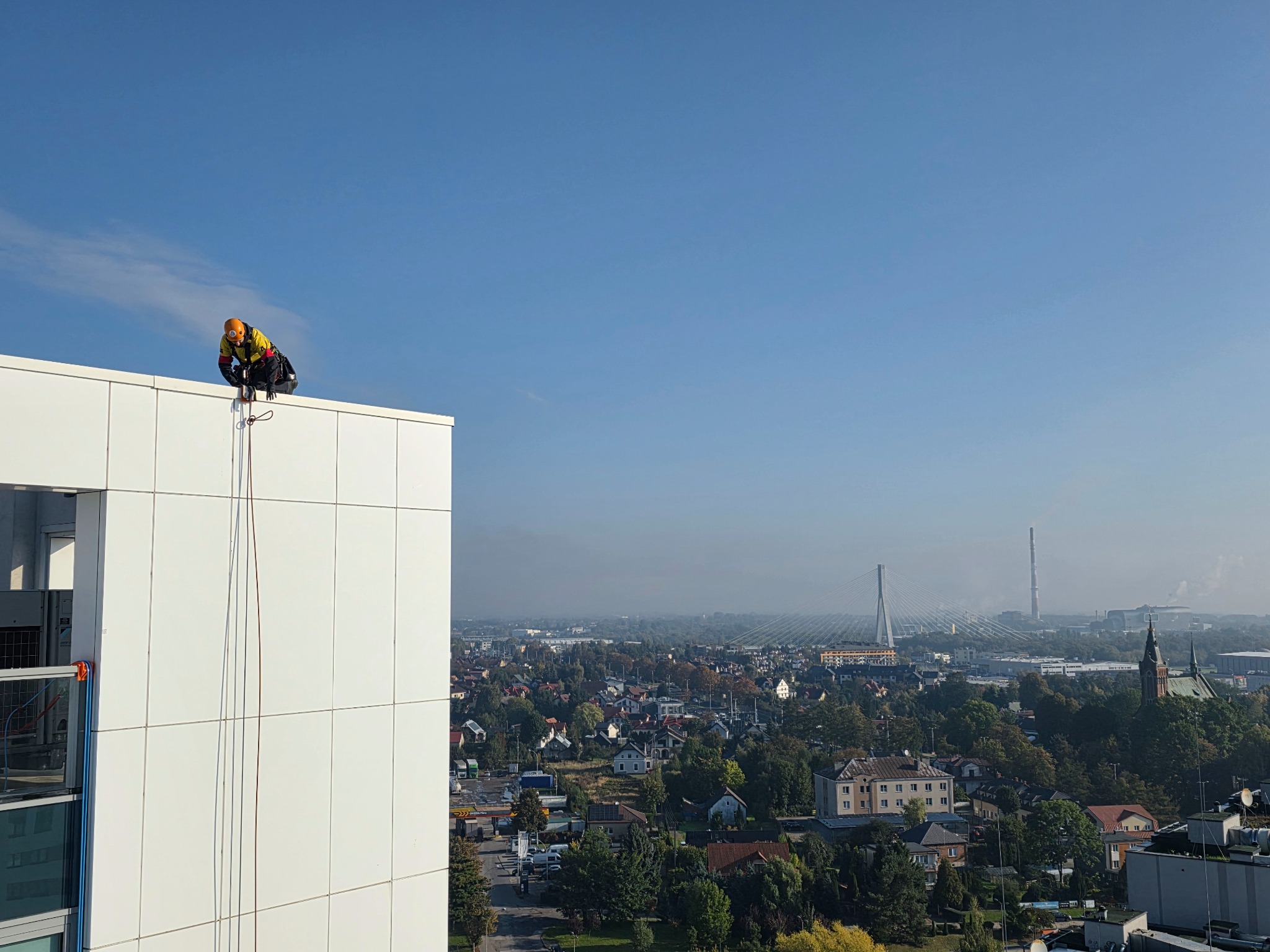 Pracownik w kasku i uprzęży na krawędzi wysokiego budynku z białymi panelami, przygotowujący się do pracy na wysokości. W tle panorama miasta z mostem i kominem.