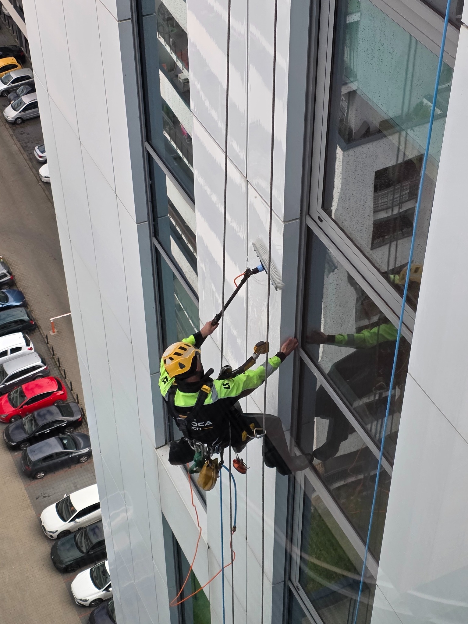 Pracownik w uprzęży, z żółtym kaskiem, czyści okna na wysokościowcu za pomocą szczotki na teleskopowym kiju. Widok z góry na ulicę z zaparkowanymi autami.