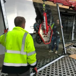 Montaż dużej szyby przez dwóch pracowników, jeden w jaskrawej kamizelce, drugi w kamizelce roboczej, przy użyciu specjalistycznego podnośnika próżniowego na tle budynku z elementami elewacji.