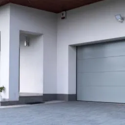 Nowoczesny dom jednorodzinny z szarą bramą garażową segmentową, widoczne okno z drewnianą ramą i fragment ogrodu z ławeczką, oświetlenie zewnętrzne.