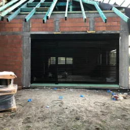 Montaż czarnej, poziomej rolety zewnętrznej na surowym budynku z widoczną konstrukcją dachu i porozrzucanymi elementami montażowymi na ziemi.