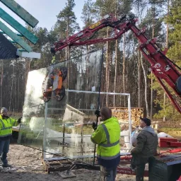 Montaż dużej szyby okiennej za pomocą dźwigu ssącego, widoczni pracownicy w odblaskowych kamizelkach, surowy stan budynku w tle las