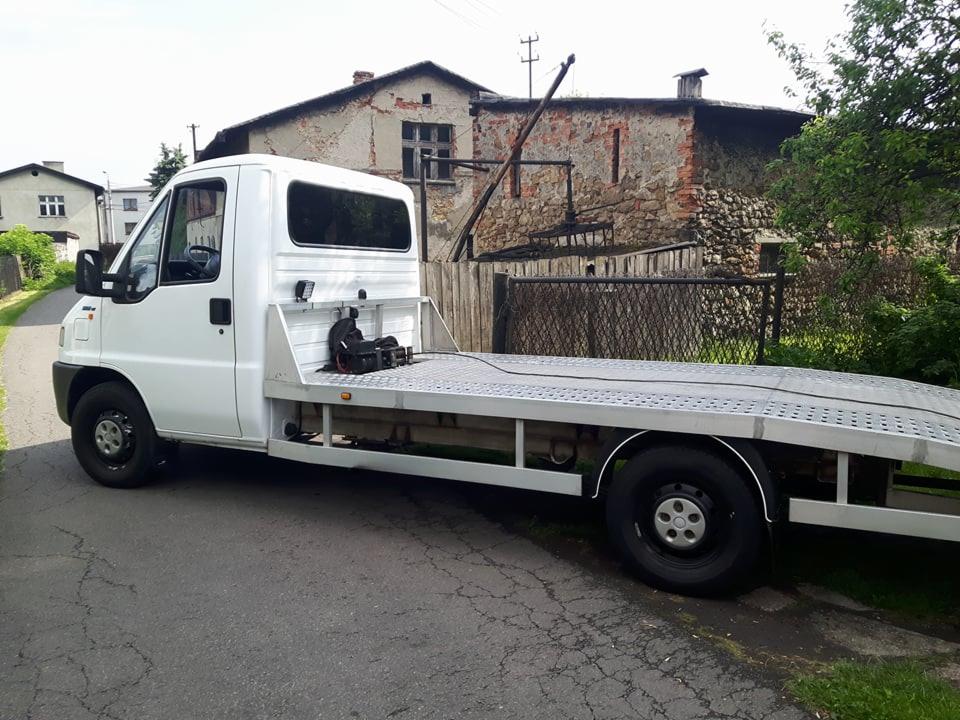 Biała laweta na tle wiejskiego krajobrazu z budynkiem w stanie surowym.