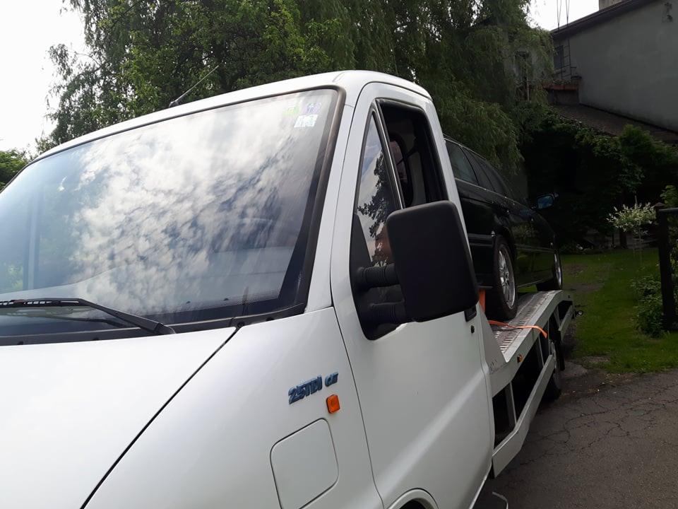 Biała laweta z załadowanym czarnym samochodem osobowym, widok z boku, tło z zielenią i budynkiem.