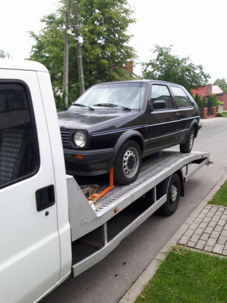 Czarny Volkswagen Golf II na srebrnej lawecie z pomarańczowymi pasami zabezpieczającymi, widok z boku na tle zieleni i zabudowań.