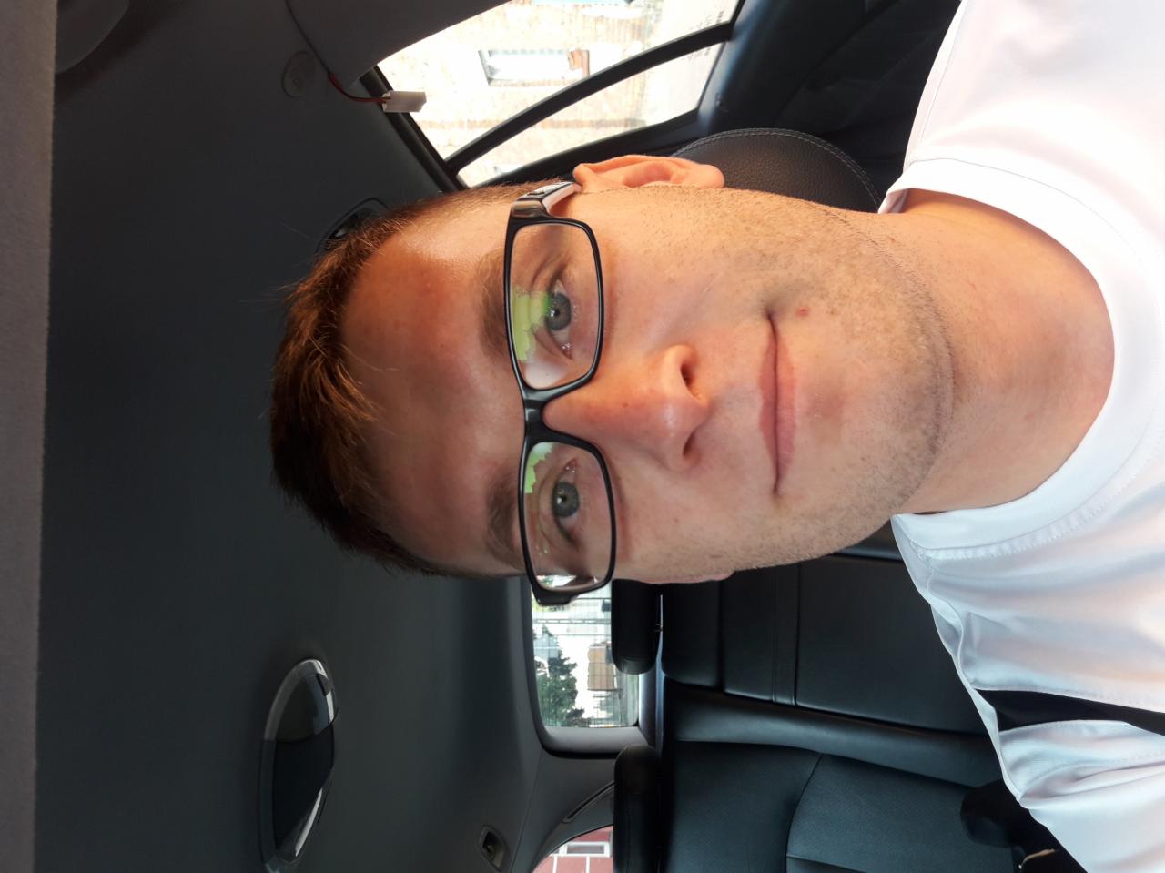 Portret mężczyzny w okularach, siedzącego na tylnym siedzeniu samochodu z czarną skórzaną tapicerką.