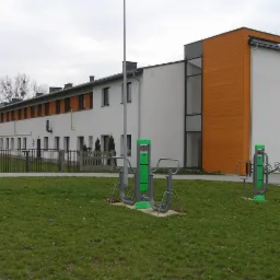 Dwukondygnacyjny budynek z białą elewacją i elementami z drewnianą okładziną, widoczny z perspektywy parku z zielonymi urządzeniami do ćwiczeń na świeżym powietrzu.