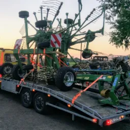 Żółta laweta przewozi zielony przetrząsacz karuzelowy Krone DRO 30/26, zabezpieczony pomarańczowymi pasami transportowymi, w tle budynek gospodarczy i drzewo liściaste.