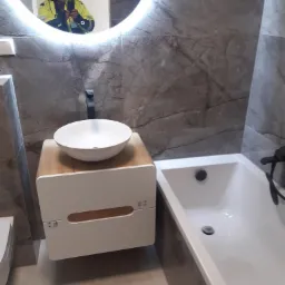 Wykończona łazienka z umywalką nablatową na szafce z drewnianymi akcentami, obok wanna, ściany wyłożone płytkami imitującymi marmur, w lustrze odbija się osoba w odblaskowej kurtce robiąca zdjęcie.