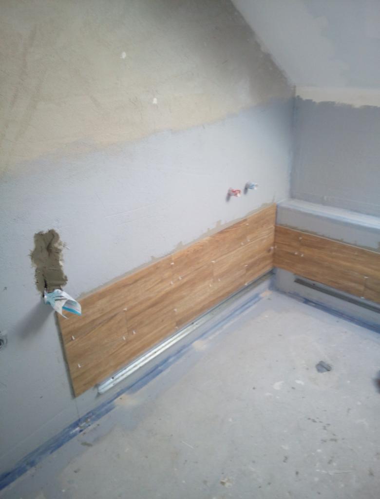 Układanie płytek imitujących drewno na ścianie w łazience, widoczna listwa poziomująca i resztki zaprawy przy otworze na rurę.