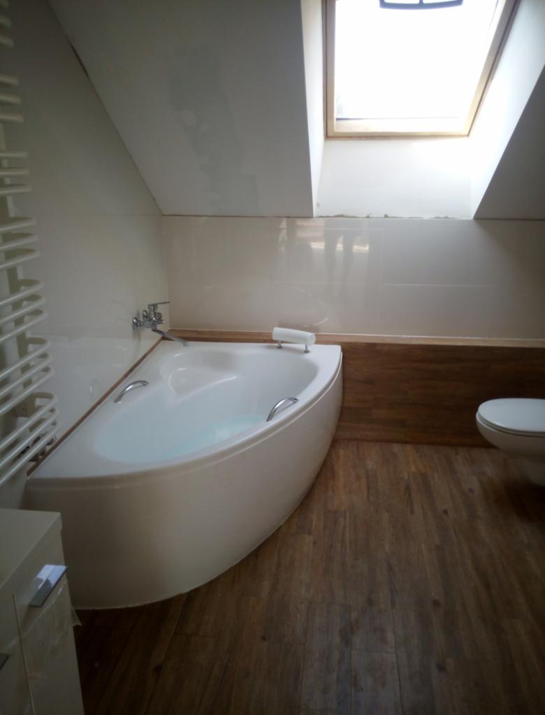 Biała wanna narożna w łazience na poddaszu z drewnopodobnymi płytkami, widoczne okno dachowe i grzejnik drabinkowy.