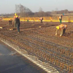 Prace zbrojarskie na budowie: widok z góry na rozległą siatkę zbrojeniową, robotnicy w odblaskowych kamizelkach i kaskach pracują nad ułożeniem zbrojenia, w tle krajobraz z drzewami i konstrukcją...