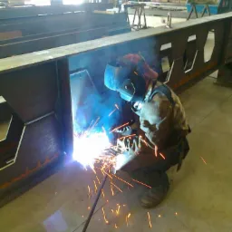Spawacz w kasku spawalniczym spawa metalową konstrukcję hali, widoczne iskry i dym spawalniczy, w tle widoczne elementy konstrukcyjne hali.