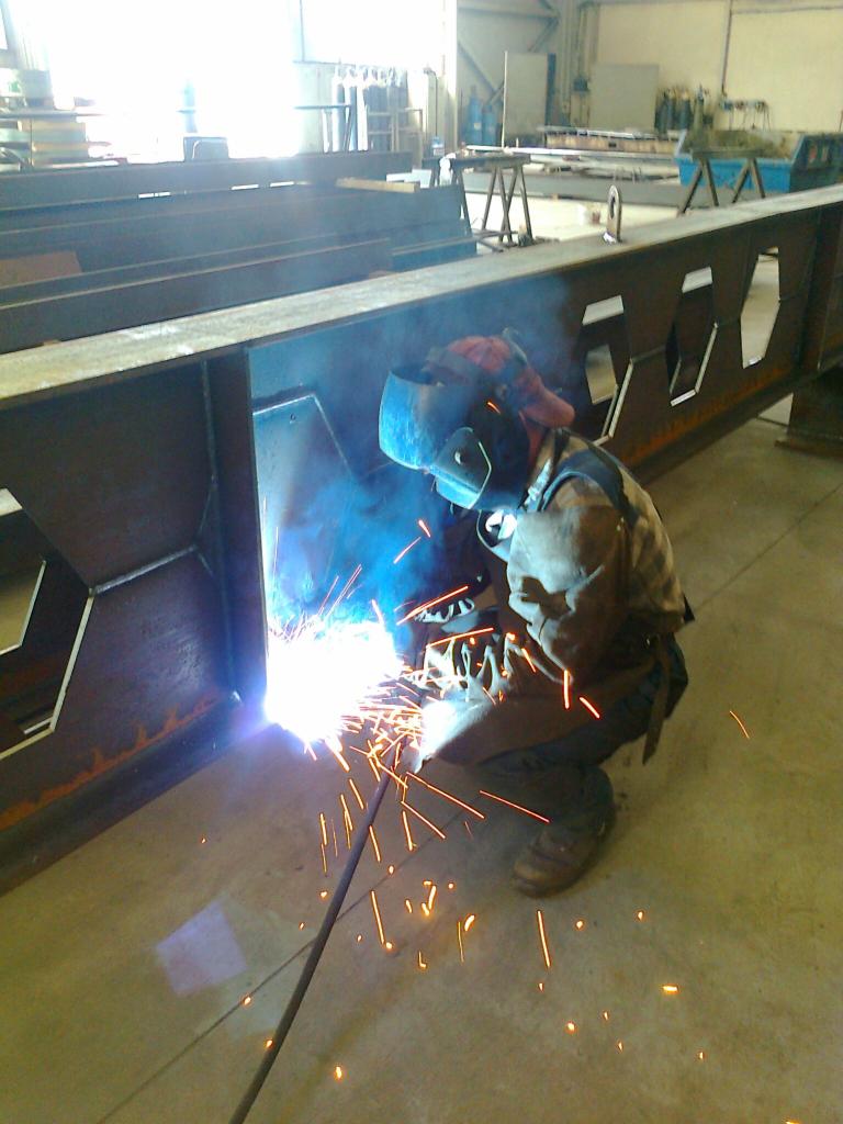 Spawacz w kasku spawalniczym spawa metalową konstrukcję hali, widoczne iskry i dym spawalniczy, w tle widoczne elementy konstrukcyjne hali.