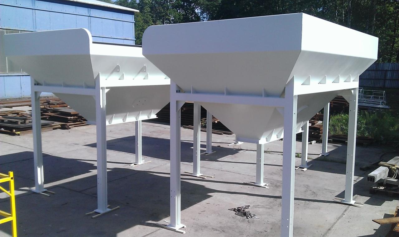 Dwa białe, stalowe zbiorniki na konstrukcji wsporczej, ustawione na betonowym placu, w tle budynek i zieleń