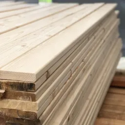 Stos jasnych, heblowanych desek z widocznym rysunkiem słojów drewna, ułożonych jedna na drugiej, z ciemniejszymi deskami na spodzie stosu.