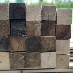 Kwadratowe kantówki z widocznymi słojami drewna, niektóre z ciemnymi przebarwieniami, ułożone w stos w Andrychowie, na tle zieleni.