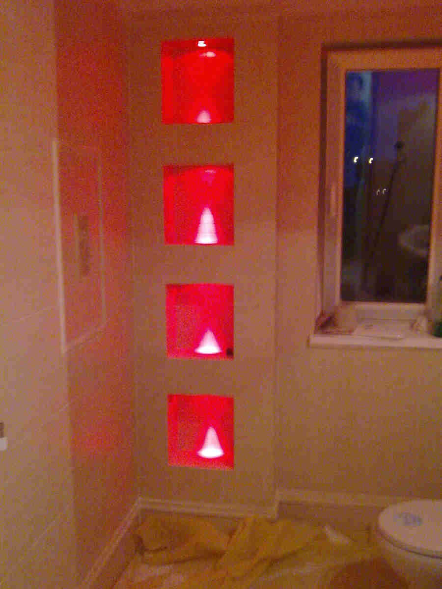 Wykończona łazienka z czterema kwadratowymi wnękami oświetlonymi czerwonym światłem w ścianie, widoczny fragment toalety i okno z parapetem.