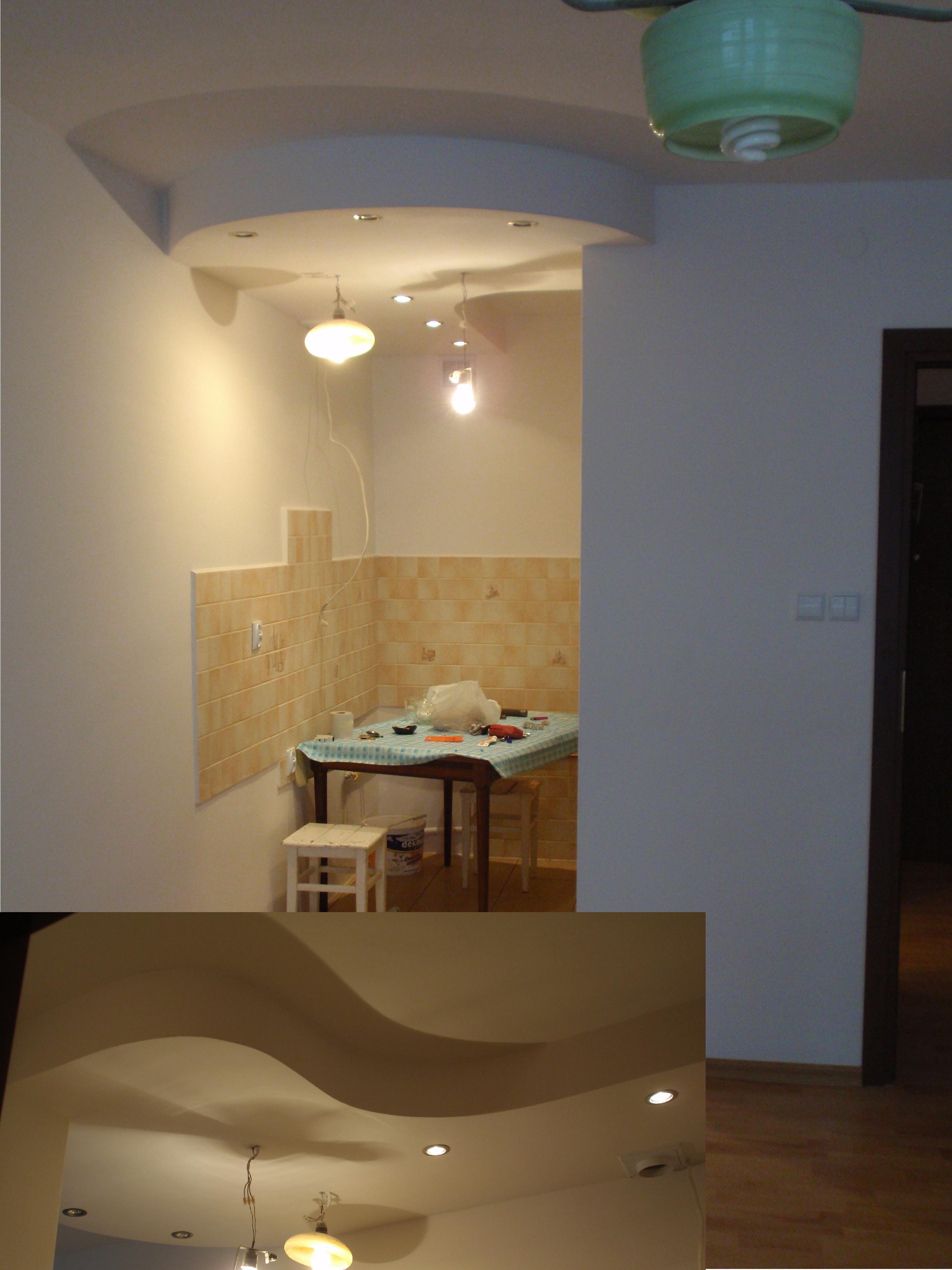 Aranżacja kuchni z widocznym fragmentem wykończonej ściany z beżowymi płytkami, stołem roboczym z narzędziami i półką sufitową z oświetleniem punktowym w tle.