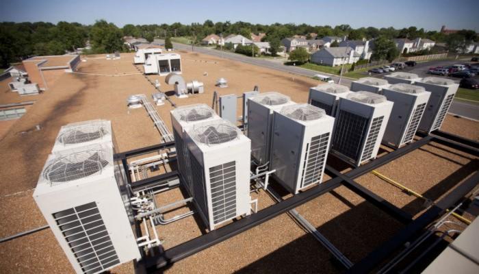 Rząd białych, przemysłowych jednostek zewnętrznych na dachu budynku, połączonych rurami i wspartych na czarnej metalowej konstrukcji, z widokiem na osiedle domów jednorodzinnych w tle.