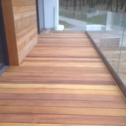 Nowoczesny taras z desek kompozytowych w odcieniach brązu, z szklaną balustradą, widok na las w tle.