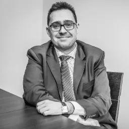 Portret mężczyzny w garniturze siedzącego przy drewnianym stole, czarno-białe zdjęcie z delikatnym uśmiechem i zegarkiem na nadgarstku.