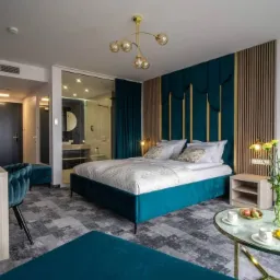 Turkusowe łóżko z tapicerowanym wezgłowiem w eleganckim pokoju hotelowym, widoczna łazienka za szklaną ścianą i stolik kawowy z deserami.