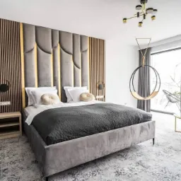 Szara sypialnia z tapicerowanym wezgłowiem z pionowymi panelami, drewniane lamele na ścianie, okrągła huśtawka pod sufitem i balkon z widokiem na zimowy krajobraz.