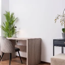 Jasny drewniany biurko z krzesłem w romby, lampka biurkowa i roślina doniczkowa, minimalistyczny wystrój wnętrza z drewnianą podłogą.