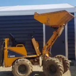 Żółta, używana mini ładowarka o udźwigu 1 tony, z podniesioną łyżką, stoi na piaszczystym terenie przed ciemną halą z białym dachem.