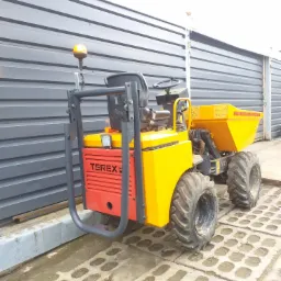 Żółto-czerwona wywrotka budowlana Terex na betonowej nawierzchni, z szarą ścianą w tle.