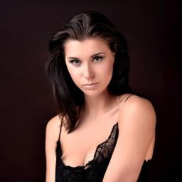 profifoto.pl - Portret młodej kobiety z ciemnymi włosami, w czarnej koronkowej bieliźnie, na ciemnym tle, z logo fotografa w prawym dolnym rogu.