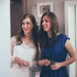 Panna młoda w koronkowej sukni i druhna w niebieskiej sukience, uśmiechnięte, stojące w drzwiach, z widocznym zdjęciem ślubnym w tle.