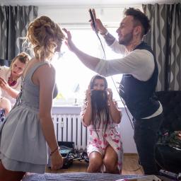 profifoto.pl - Przygotowania panny młodej: fryzjer stylizuje włosy prostownicą, makijażystka poprawia makijaż, a koleżanka trzyma włosy przed twarzą, udając brodę.