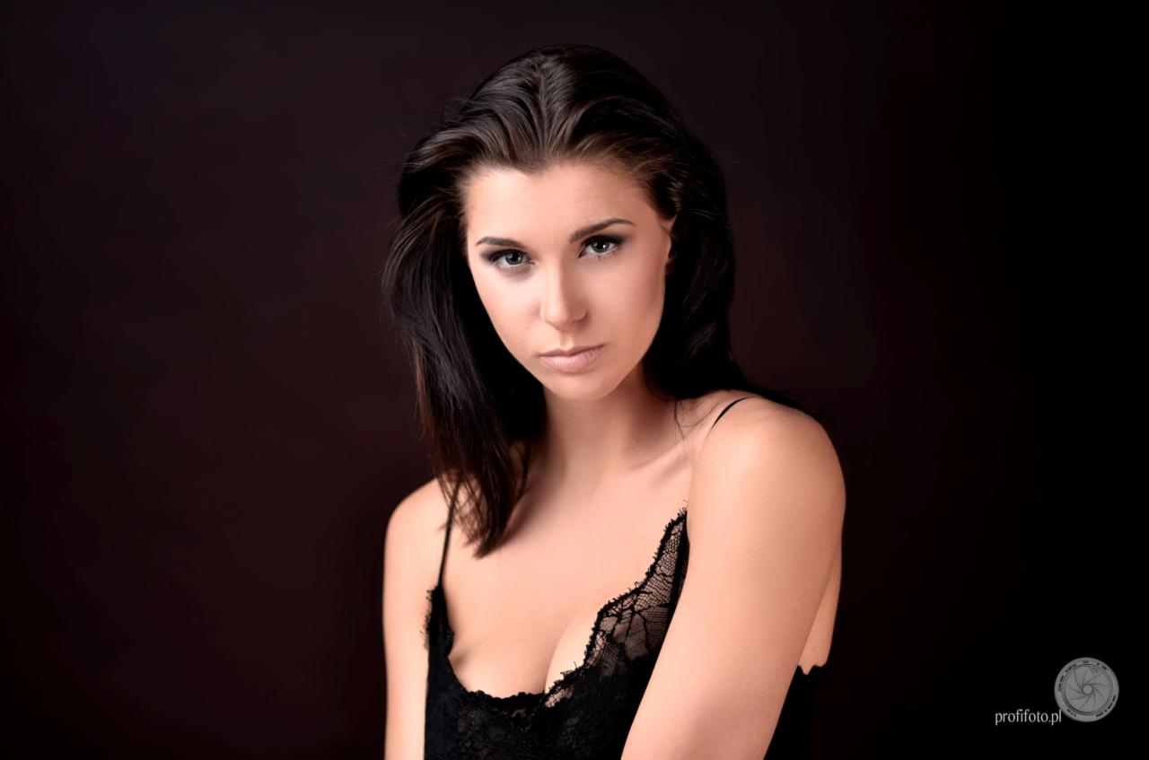 Portret młodej kobiety z ciemnymi włosami, w czarnej koronkowej bieliźnie, na ciemnym tle, z logo fotografa w prawym dolnym rogu.