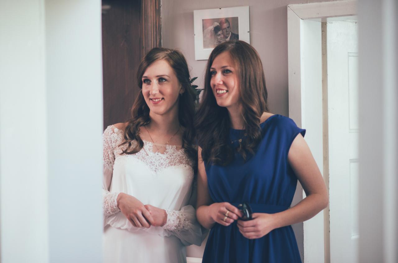 Panna młoda w koronkowej sukni i druhna w niebieskiej sukience, uśmiechnięte, stojące w drzwiach, z widocznym zdjęciem ślubnym w tle.
