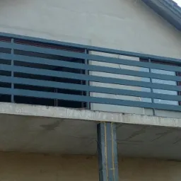 Szara, pozioma balustrada balkonowa z prostokątnych profili, zamontowana na surowym, betonowym balkonie domu jednorodzinnego w trakcie budowy.